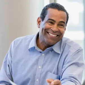 A man wearing a light blue button-up shirt sits at a table, smiling warmly and looking slightly to the side, as if discussing netsuite solutions or how to hire netsuite experts, with a blurred background behind him.