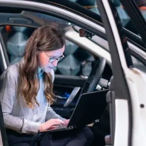 A woman with long brown hair and glasses sits in the driver's seat of a car, working on a laptop with a focused expression. She appears to be managing tasks in NetSuite, dressed in a light-colored blouse.